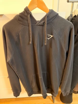 Svart hoodie från Gymshark - Säljer en svart hoodie från Gymshark med klassisk huva och dragsnören. Hoodien har en ficka framtill och Gymshark-logga på bröstet. Tillverkad i mjukt material som passar perfekt till chill eller träning. Crest 