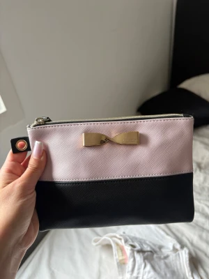 Victoria's Secret necesär  - Snygg clutch från Victoria's Secret i svart och ljusrosa med guldfärgad rosettdetalj framtill. Väskan har dragkedja upptill och är tillverkad i ett tåligt, strukturerat material. Perfekt storlek för det viktigaste och enkel att bära med sig.