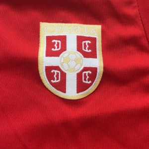 Serbien Matic 21 fotbollströja röd - Röd fotbollströja från Serbiens landslag med vitt tryck, nummer 21 och namnet Matic på ryggen. Tröjan har korta ärmar med vita detaljer längs axlar och ärmslut samt Serbiens emblem på bröstet. Tillverkad i lätt och ventilerande polyester.