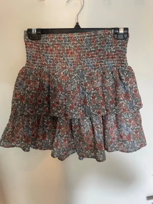 Blommig volangkjol från SRN - Supersöt kort blommig kjol från Ullared med volanger och smock i midjan. Kjolen har ett färgglatt mönster i rött, blått och vitt och är perfekt för dig som gillar en lekfull och trendig stil. Lätt och luftig, passar perfekt till varma dagar.