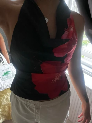 Svart halterneck topp med röda blommor - Snygg svart halterneck topp med stora röda blommor. Toppen har en draperad halsringning och är ärmlös, vilket ger en trendig och somrig vibe. Perfekt att styla med både jeans och kjol för en cool look.