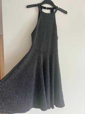 Svart glittrig kortklänning - Supersnygg svart kortklänning med glittrigt tyg och hög hals. Klänningen har ärmlös design och ett lätt utställt kjolparti som ger en fin siluett. Perfekt för dig som vill sticka ut med lite extra glitter.