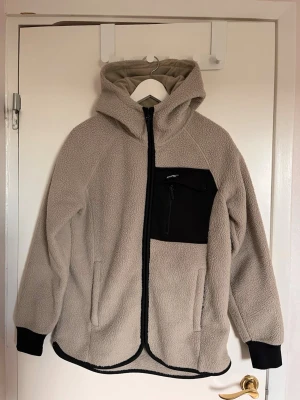 Beige teddyjacka med huva - Mysig beige teddyjacka med huva och hel dragkedja framtill. Jackan har svart bröstficka med dragkedja, svarta muddar vid ärmsluten och svarta detaljer längs kanterna. Perfekt för kalla dagar och riktigt skön att bära.