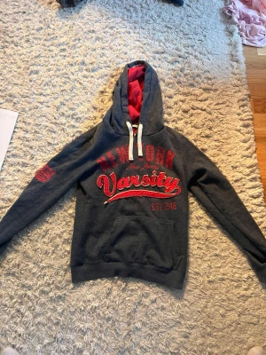 Mörkgrå hoodie New York Varsity - Mörkgrå hoodie med röd insida i huvan och stora röda tryck på bröstet där det står 'New York Varsity'. Känguruficka framtill och vita dragsnören i huvan. Mjuk och skön, perfekt för chill dagar. Märkt Luxury Fashion. Storlek S men passar även XS!!