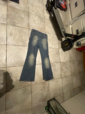 Blå bootcut jeans med slitningar - Säljer ett par blå bootcut jeans med tydliga slitningar och ljusa partier på framsidan och baksidan av benen. Jeansen har klassiska fem fickor och normal midja. Perfekta för en avslappnad och trendig look.