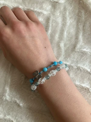 Två pärlarmband med fjäril - Säljer två armband med pärlor, ett i blått med en silverfärgad fjärilsberlock och ett i vitt och turkost med genomskinliga pärlor med mönster 😊