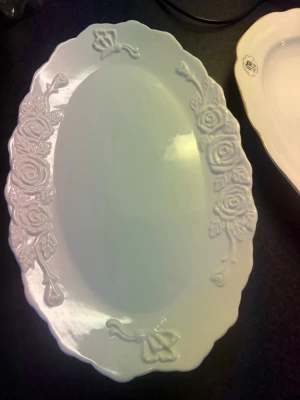 Vitt ovalt serveringsfat med rosor - Säljer ett vitt ovalt serveringsfat i porslin med upphöjt rosmönster längs kanterna. Fatet har vågiga kanter och en klassisk, elegant design som passar perfekt till middagsbjudningar eller som dekoration.