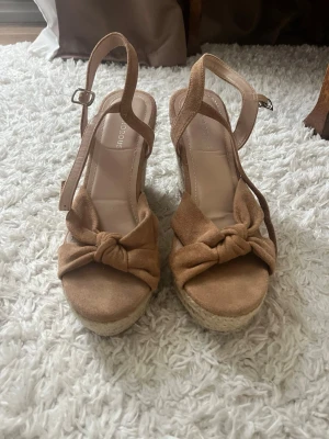 Beiga kilklackade sandaler med rosett - Säljer ett par snygga beiga sandaler med kilklack och flätad sula. Fint skick och använda 1 gång 