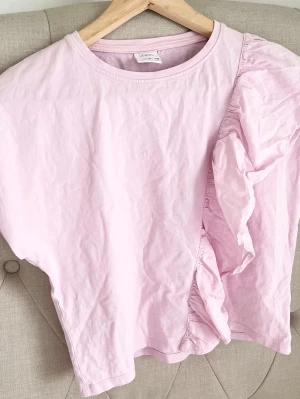Ljusrosa t-shirt med volangdetalj - Säljer en ljusrosa t-shirt med rund hals och korta ärmar. T-shirten har en snygg volangdetalj som går diagonalt över framsidan och ger en unik look. Perfekt för dig som vill ha något extra i garderoben.