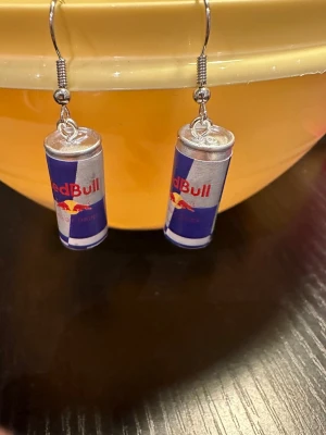 Örhängen med Red Bull-burkar - Unika örhängen formade som små Red Bull-burkar i silver, blått och rött. Perfekta för dig som vill sticka ut med en lekfull och kreativ accessoar. Krokar i metall och detaljerad design som liknar riktiga energidrycksburkar.