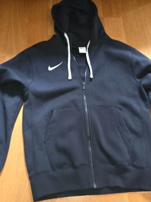 Svart zip hoodie från Nike - Säljer en svart hoodie från Nike med dragkedja framtill och vita snören i huvan. Klassisk Nike-logga på bröstet och stora fickor framtill. Perfekt för en sportig och avslappnad stil. Materialet är mjukt och bekvämt, troligen bomullsblandning.