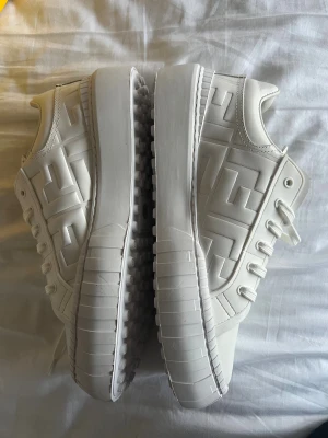 Vita Fendi sneakers med FF-mönster - Kommer med kartongen. Säljer ett par vita Fendi sneakers i skinn med präglat FF-mönster på sidorna. Skorna har rund tå, platt sula med gummidetaljer undertill och klassisk snörning. Snygg och clean design som sticker ut med Fendis ikoniska logga. 