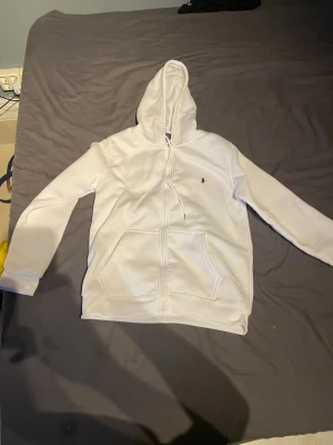 Vit hoodie med dragkedja från Polo Ralph Lauren - Sprillans ny Snygg vit hoodie från Polo Ralph Lauren med dragkedja framtill och klassisk huva. Har två stora fickor och den ikoniska broderade loggan på bröstet. Perfekt för en clean och stilren look. Mjuk och skön bomullskänsla.