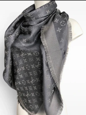 Grå Louis Vuitton sjal med monogram - Snygg grå sjal från Louis Vuitton med det klassiska monogrammönstret i ton-i-ton. Sjalen har fransade kanter och är tillverkad i ett mjukt, glansigt material som ger en lyxig känsla. Perfekt att svepa runt halsen för en trendig look.