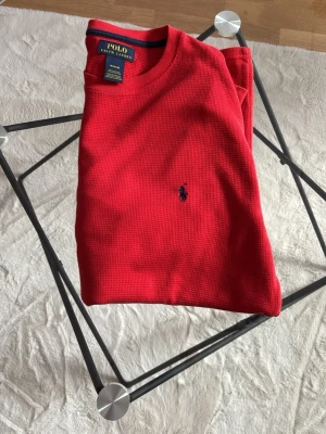 Röd t-shirt från Polo Ralph Lauren - Snygg röd t-shirt från Polo Ralph Lauren med klassisk blå logga broderad på bröstet. T-shirten har rund hals, korta ärmar och är tillverkad i ett strukturerat bomullstyg som ger en skön känsla. Perfekt till jeans eller shorts.