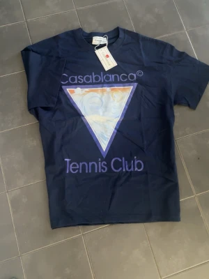 Mörkblå Casablanca Tennis Club t-shirt - Snygg mörkblå t-shirt från Casablanca med stort tryck framtill där det står 'Casablanca Tennis Club' och en färgglad triangel. Klassisk passform och rund hals. Perfekt för dig som gillar streetwear och sportig stil.