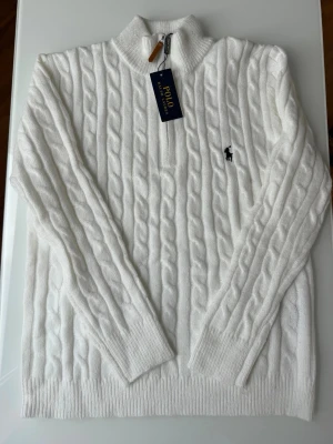 Vit kabelstickad tröja Polo Ralph Lauren - Vit kabelstickad tröja från Polo Ralph Lauren med halv dragkedja och hög krage. Tröjan har det klassiska broderade logotypen på bröstet och ribbade muddar vid ärmslut och nederkant. Perfekt för en stilren och tidlös look.
