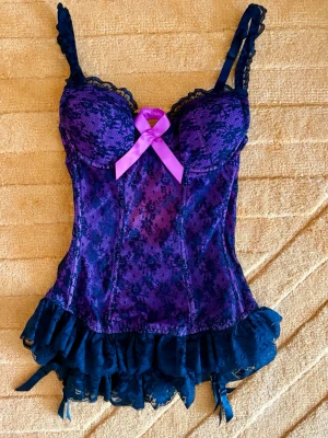 Lila spetskorsett med volang och rosett - Snygg lila korsett i spets med svarta detaljer och volangkant nedtill. Utrustad med reglerbara axelband, vadderade kupor och en dekorativ lila rosett framtill. Materialet är genomskinlig spets och mesh, vilket ger en feminin och trendig look.