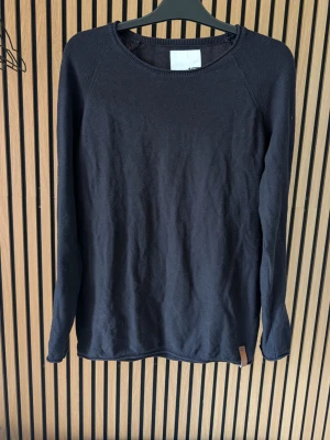 Svart stickad tröja med rund hals - En enkel och stilren svart stickad tröja med rund halsringning och långa ärmar. Tröjan har en diskret etikett nertill och är tillverkad i ett mjukt, tunt material som passar perfekt till lager på lager. Snygg och tidlös modell som funkar till det mesta.