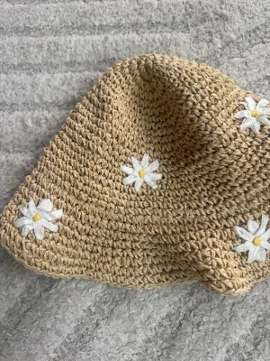 Beige virkad solhatt med prästkragar - Söt beige virkad solhatt från H&M, dekorerad med vita prästkragar med gula mittpunkter. Hatten har en mjuk och böjlig form och är perfekt för soliga dagar. Tillverkad i ett flätat material som ger en naturlig och somrig känsla.