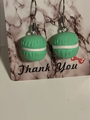Gröna macaron örhängen - Säljer ett par unika örhängen formade som gröna macarons med vit fyllning. De har en söt och lekfull design och är tillverkade i plast med metallkrokar. Perfekta för dig som vill sticka ut med något annorlunda och färgglatt. Örhängens krokarna är i rostfritt stål.