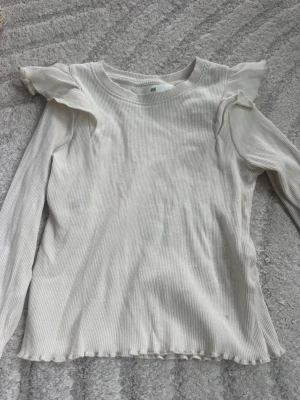 Vit långärmad topp med volang Zara - Söt vit långärmad topp från Zara i ribbat material. Toppen har volangdetaljer vid axlarna och en lätt rynkad kant nedtill. Perfekt för dig som gillar en enkel men ändå trendig stil.
