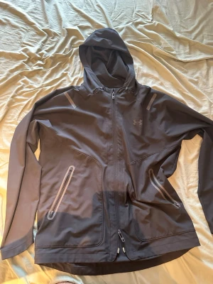 Under Armour jacka - Snygg svart vindjacka från Under Armour med huva och hel dragkedja. Jackan har två dragkedjeförsedda fickor framtill och diskreta reflexdetaljer på axlarna. Tillverkad i ett lätt och vindtätt material, perfekt för blåsiga dagar.