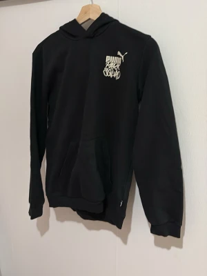 Svart hoodie från Puma med tryck - Säljer en svart hoodie från Puma med vit logga och tryck på bröstet. Hoodien har huva, känguruficka och ribbade muddar vid ärmslut och nederkant. Perfekt för en sportig och avslappnad stil.