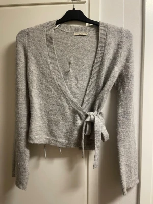 Grå omlottkofta med knytband - Mysig grå omlottkofta med långa ärmar och knytband i midjan. Koftan har en djup v-ringning och ribbade kanter. Perfekt att slänga över en topp för en avslappnad look.
