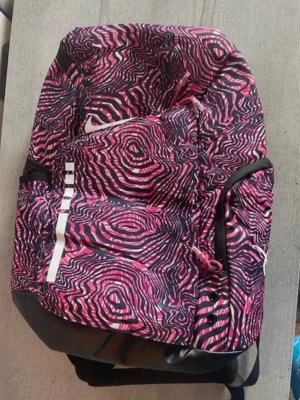 Nike Elite ryggsäck rosa mönster - Nike Elite ryggsäck med ett unikt rosa och svart mönster som påminner om träådring. Ryggsäcken har flera fack, vadderade axelband med ELITE-tryck och sidofickor i mesh. Perfekt för skolan eller träningen och rymmer mycket. Tillverkad i slitstarkt syntetmaterial. Helt ny.