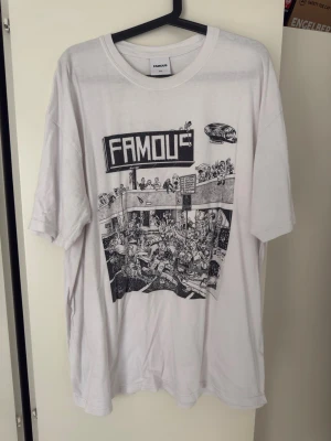 Vit oversized t-shirt från Famous - Vit oversized t-shirt från Famous med svart grafiskt tryck framtill som föreställer en tecknad scen och stor text 'FAMOUS' på bröstet. Klassisk rund hals och korta ärmar. Perfekt för dig som gillar streetwear och statement-plagg.