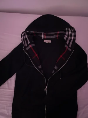 Svart hoodie med rutigt foder - Pris går att diskutera!!! Säljer en svart hoodie med dragkedja och huva. Insidan av huvan har ett klassiskt rutigt mönster i rött, vitt och grått. Hoodien har fickor framtill och snörning i huvan. Perfekt för en chill och stilren look.