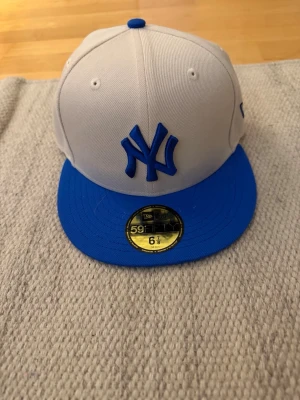 Vit och blå New Era Yankees keps - Snygg vit keps från New Era med blått New York Yankees-logo framtill och blå skärm. Klassisk 59Fifty-modell med rak skärm och broderade detaljer. Perfekt för dig som vill ha en stilren och sportig look.