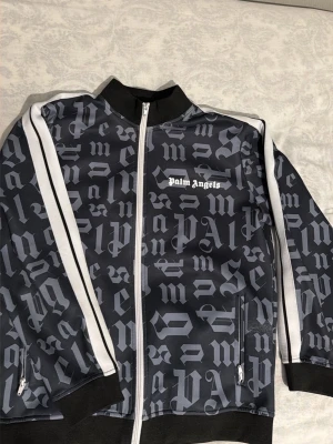 Palm Angels svart ziptröja - Palm Angels ziptröja i svart med vit logga på bröstet och gotiskt mönster över hela plagget. Tröjan har vita ränder längs ärmarna, hög krage och muddar vid ärmslut och nederkant. Perfekt för en streetwear-look.