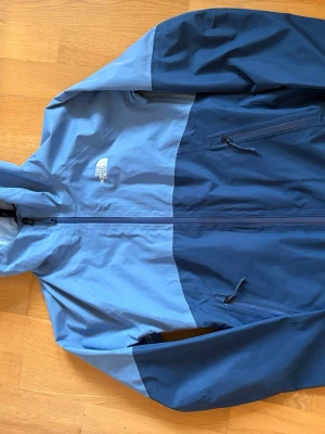 Blå vindjacka från The North Face - Snygg tvåfärgad blå vindjacka från The North Face med huva och dragkedja framtill. Jackan har två sidofickor med dragkedja och logga på bröstet och ryggen. Tillverkad i lätt och tåligt material, perfekt för blåsiga dagar. Den är i mycket bra skick! Hör av för fler bilder och prisförslag. 🤗