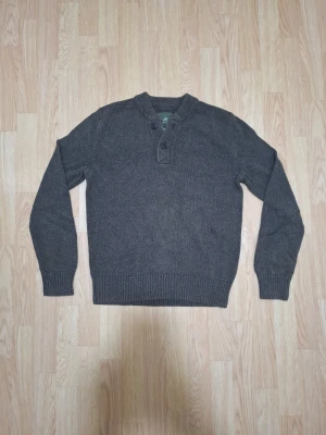 Mörkgrå stickad tröja med knappar - Mörkgrå stickad tröja med två knappar vid halsen och ribbade muddar vid ärmslut och nederkant. Tröjan har en enkel och stilren design med lång ärm och rundad halsringning. Perfekt för kyliga dagar.