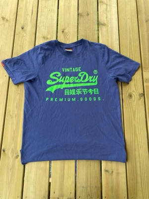 Blå Superdry t-shirt med grön text - Snygg blå t-shirt från Superdry med stort grönt tryck på bröstet och texten 'Vintage Superdry Premium Goods' samt japanska tecken. Klassisk passform och rund hals. Perfekt för dig som gillar streetwear och vill sticka ut lite extra.