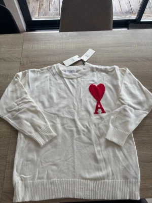 Ami paris sweatshirt - Säljer en vit stickad tröja med ribbade muddar och nederkant. På bröstet finns ett stort rött hjärta och bokstaven A i samma färg. Tröjan har rund halsringning och är gjord i mjukt material, perfekt för en trendig och avslappnad look. Hur skön som helst!