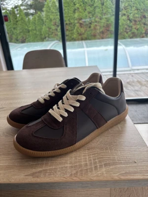 Maison margiela gats - Snygga sneakers i mörkbrunt läder och mocka med beige snörning och gummisula. Klassisk låg modell med retrovibbar och kontrasterande materialval som ger en stilren look. Perfekta för dig som gillar minimalistisk och tidlös design. defekt där bak på skorna (bild 4) därav priset är så lågt. Strlk 42 men passar även 42,5