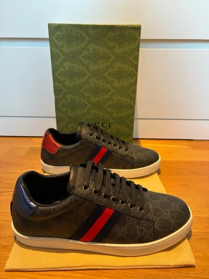 Gucci Ace sneakers  - Snygga svarta Gucci sneakers med diskret GG-mönster, klassisk röd och blå rand på sidan samt kontrastdetalj i rött på hälen. Skorna har vita sulor och svarta skosnören. Perfekta för dig som vill ha en lyxig och trendig look.