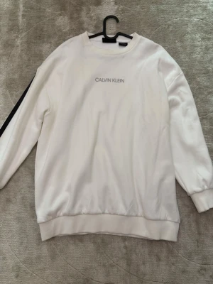 Vit sweatshirt från Calvin Klein - Vit sweatshirt från Calvin Klein med rund halsringning och diskret logga på bröstet. Tröjan har långa ärmar, ribbade muddar och en svart detalj längs ena ärmen. Perfekt för en clean och stilren look.
