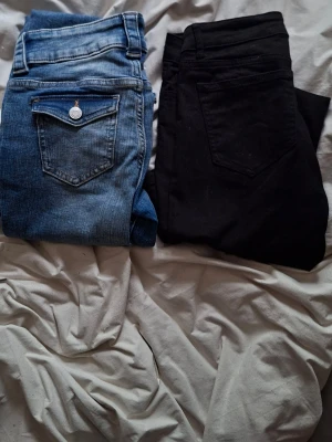 Blå och svarta jeans, utsvängda ben - Två par jeans, ett par klassiska blå jeans med ficklock och silverknapp samt ett par svarta jeans. Båda har utsvängda ben och låg midja. Perfekta till vardagslooken och funkar till det mesta. Materialet är denim, bomull, polyester och elastan. De blåa jeansen är från H&M och de svarta är från Lindex.
