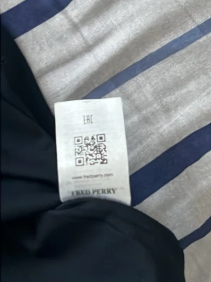 Svart t-shirt från Fred Perry - Snygg svart t-shirt från Fred Perry med klassisk rund hals och röd broderad logga på bröstet. Tillverkad i mjuk bomull för skön känsla och enkel stil. Perfekt till jeans eller shorts för en clean look.