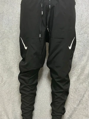 Svarta Nike träningsbyxor - Säljer ett par svarta träningsbyxor från Nike med vita loggor på framsidan av låren. Byxorna har smal passform, dragsko i midjan och är tillverkade i ett lätt och stretchigt material som passar perfekt för sport och träning.
