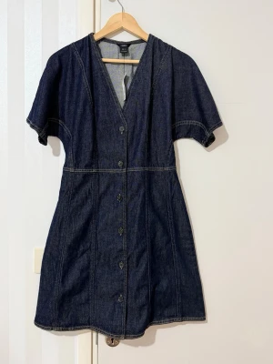 Mörkblå jeansklänning med knappar - Kort jeansklänning i mörkblå denim med v-ringning och knappar framtill. Klänningen har korta ärmar och markerad midja för en snygg siluett. Perfekt till sneakers eller boots för en cool vardagslook.