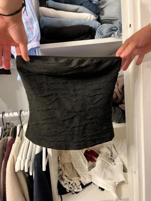 Svart strukturerad tubtopp - Säljer en svart tubtopp med snyggt strukturerat tyg. Toppen är axelbandslös och har en lätt vågig yta som ger en cool look. Perfekt att styla med jeans!