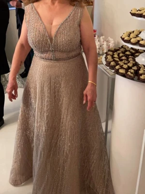 Glittrig långklänning med v-ringning - Elegant långklänning i beige med glittriga detaljer och djup v-ringning. Klänningen är ärmlös och har markerad midja samt ett lätt utställd kjol. Perfekt för speciella tillfällen där du vill glänsa.