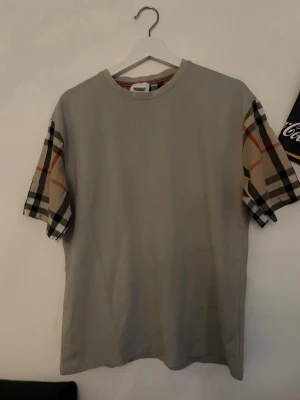 Beige t-shirt med rutiga ärmar - Snygg beige t-shirt med klassiska rutiga ärmar i beige, svart och rött. T-shirten har rund hals och normal passform. Perfekt för dig som vill ha en stilren men ändå unik look.