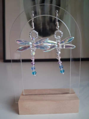 trollsländeörhängen med pärlor - Unika örhängen formade som trollsländor med skimrande vingar och dekorerade med små pärlor i vitt, lila,rosa och blått. Hängande design med silverfärgade krokar och detaljerade pärlsträngar som ger en lekfull och färgstark look. Öronkroken är nickelsäker 