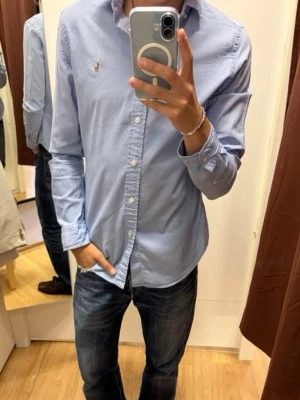 Ljusblå skjorta från Polo Ralph Lauren - Klassisk ljusblå skjorta från Polo Ralph Lauren med broderad logga på bröstet. Skjortan har lång ärm, knappar framtill och en tidlös design i bomull. Perfekt för en clean och stilren look.